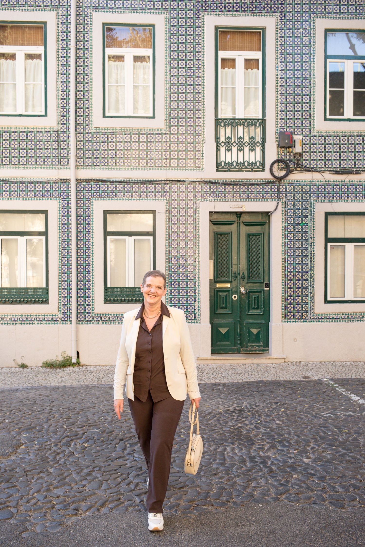 Smiling woman in a cream blazer and brown outfit walks past a blue-green tiled building with green doors on a cobblestone street.