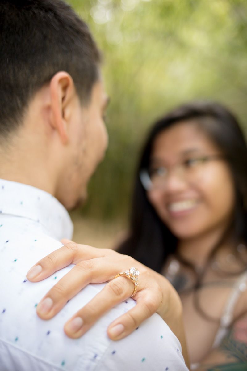 Smiling woman with glasses rests her hand on mans shoulder showing off diamond engagement ring outdoors.