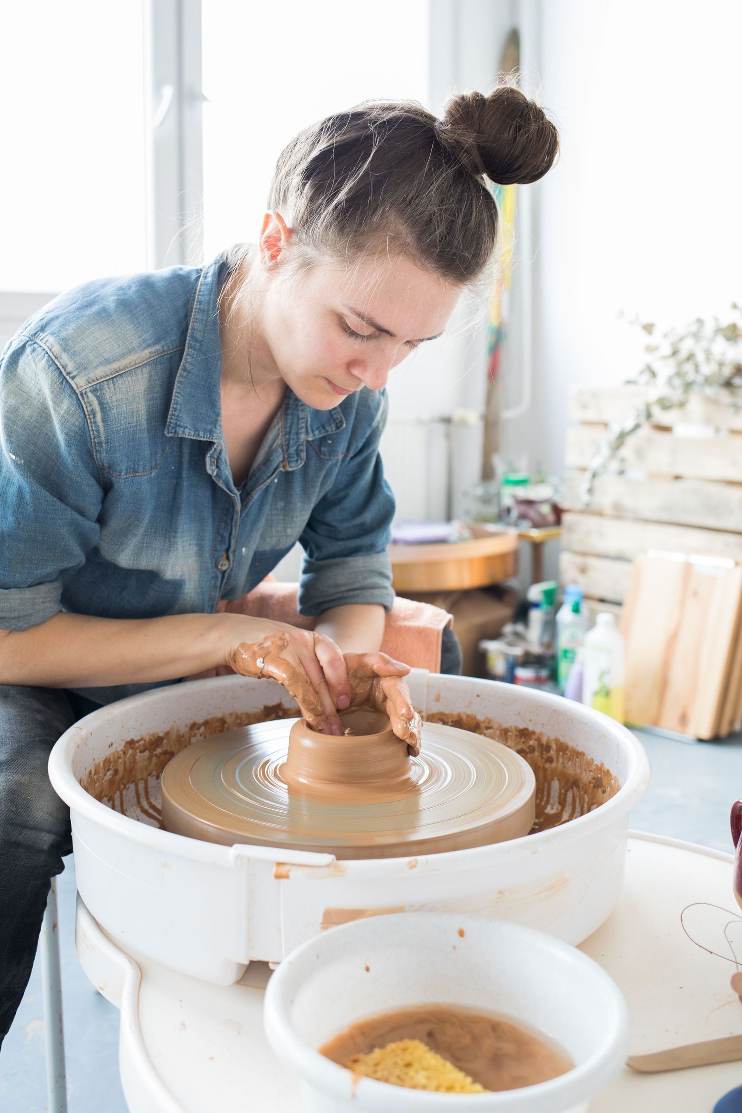A focused potter with her hair in a bun shapes wet clay on a pottery wheel in a bright, sunlit ceramics studio.