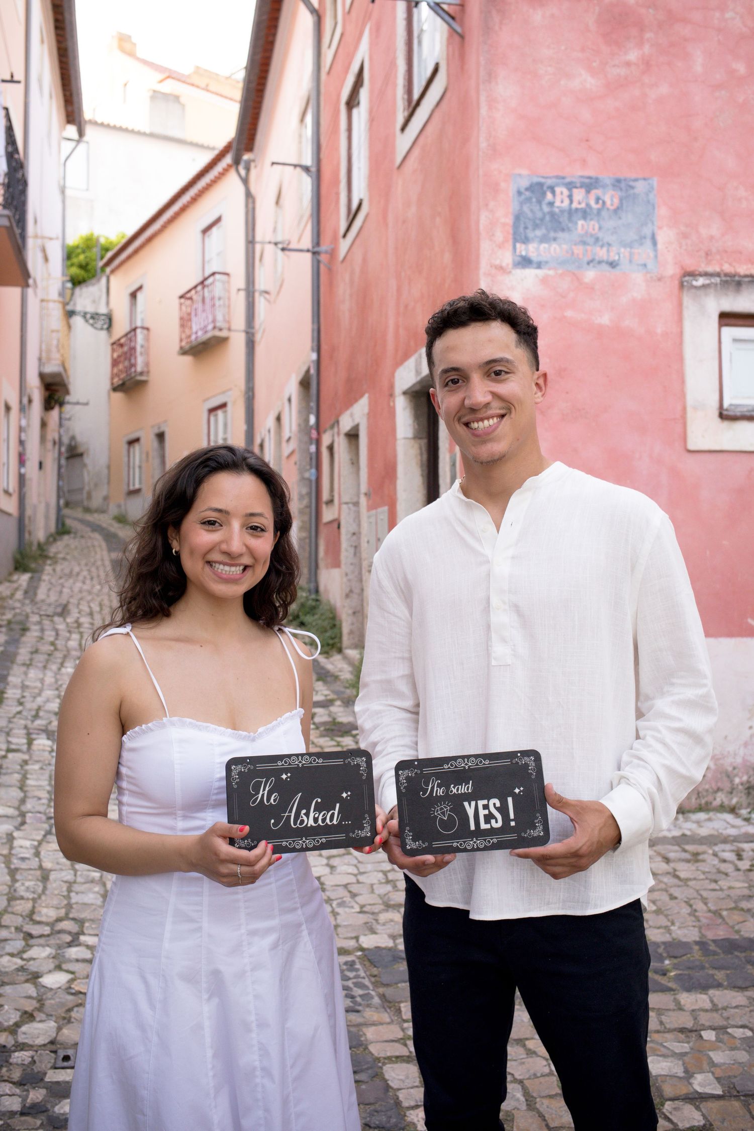 Smiling couple holding chalkboard signs reading He Asked and She Said Yes on cobblestone street.