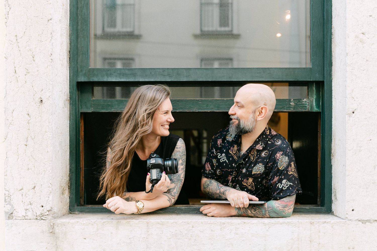 A tattooed couple laughs together leaning on a windowsill, a female photographer in Lisbon holding a camera, man in floral shirt.