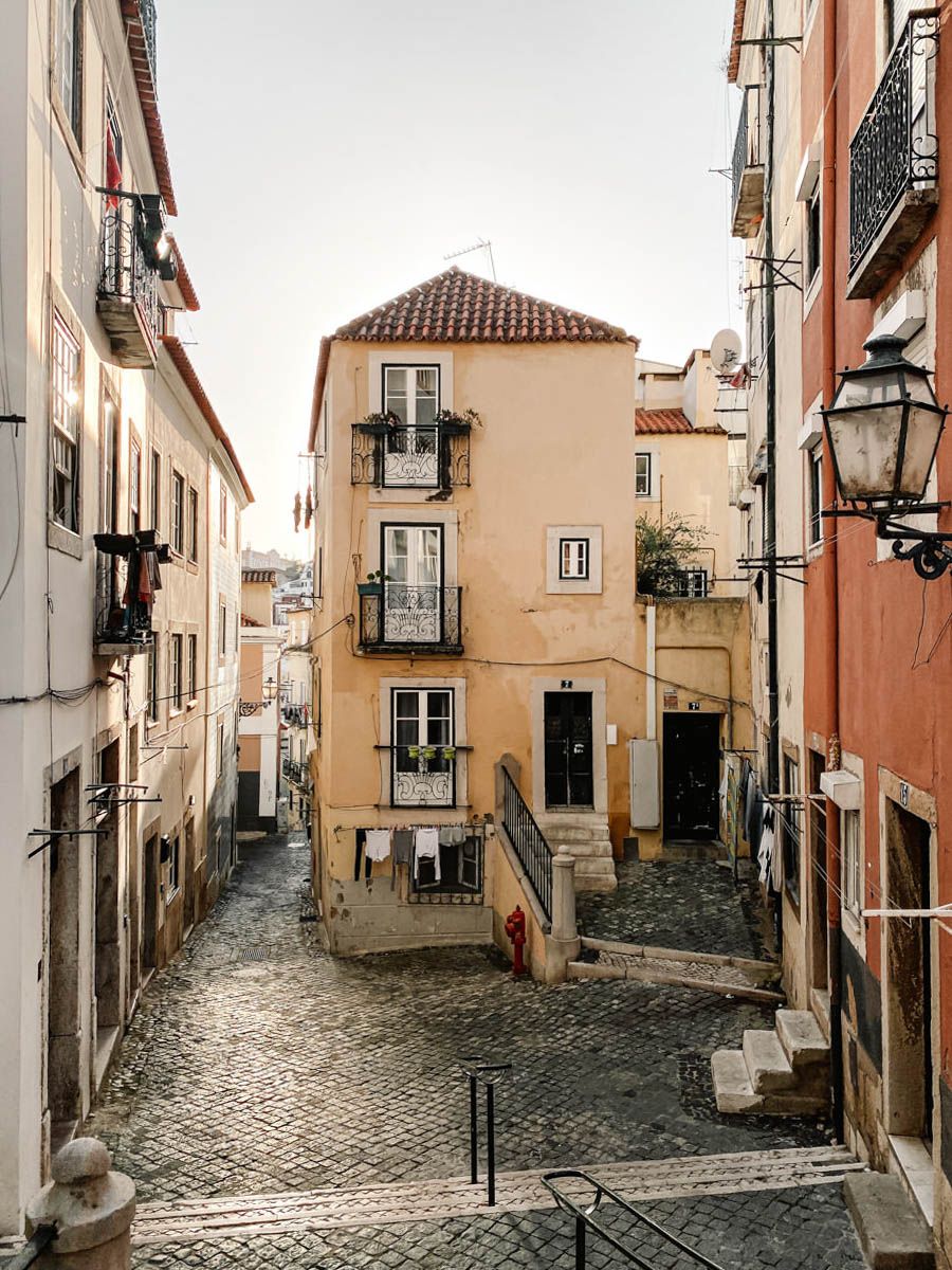 Narrow cobblestone alley in Lisbon with colorful buildings, iron railings, and a vintage street lamp in the Alfama district.