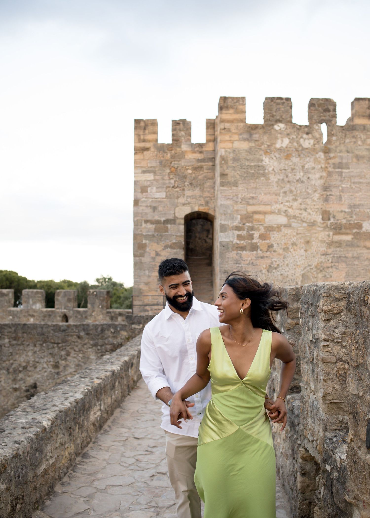 Couple holding hands and smiling while walking along ancient stone castle battlements, woman in green dress, man in white shirt.