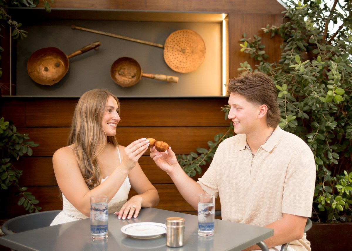 A smiling couple shares a bite of food at an outdoor patio table with copper wall decor and lush greenery behind them.