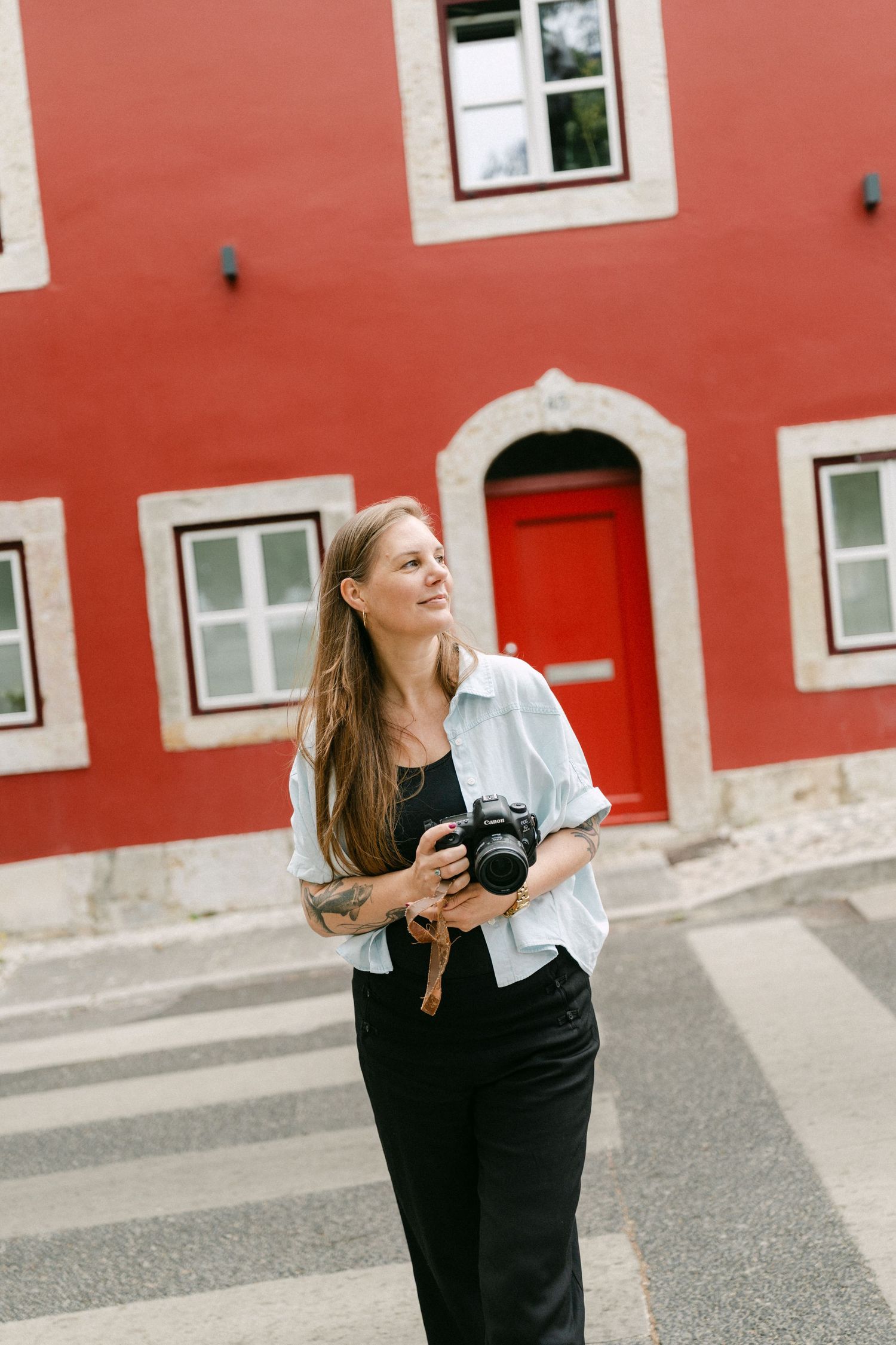 Woman photographer in Lisbon holding DSLR camera smiling outdoors near red building with arched door in Lisbon city street.