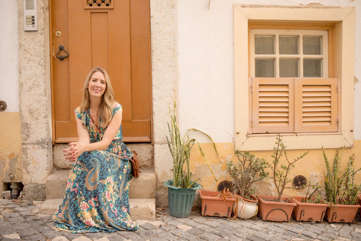 Photoshoot in Lisbon of a woman in a floral dress seated by an ochre door and shuttered window on a cobbled street.