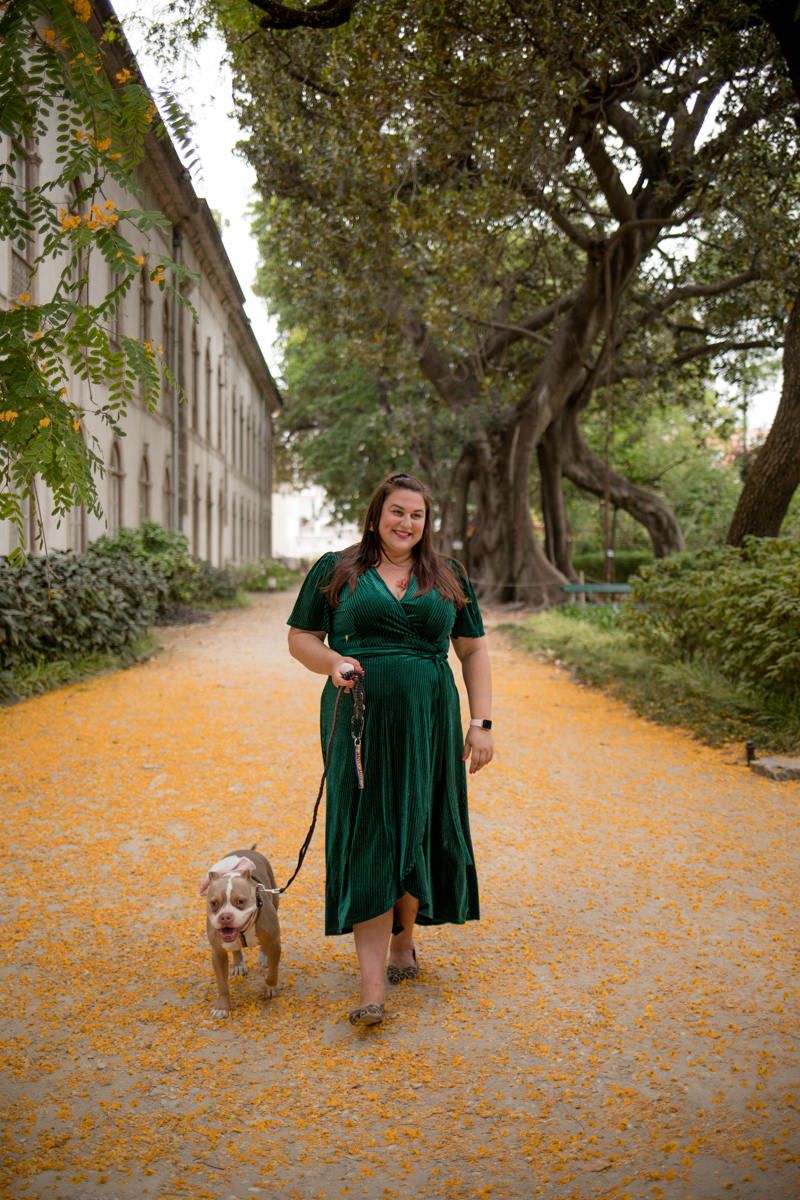 Plus-size woman in emerald green wrap dress walks dog on golden leaf-covered path near historic building.
