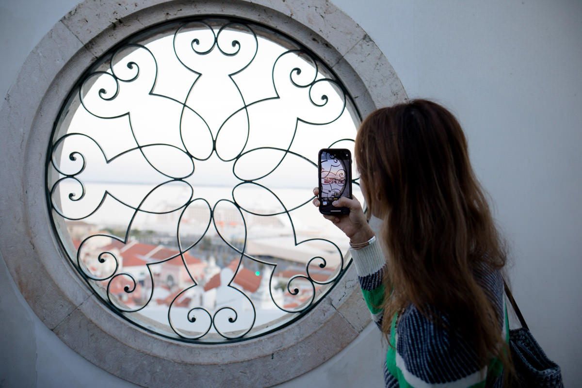 Woman photographing ornate circular window with iron scrollwork design using smartphone.