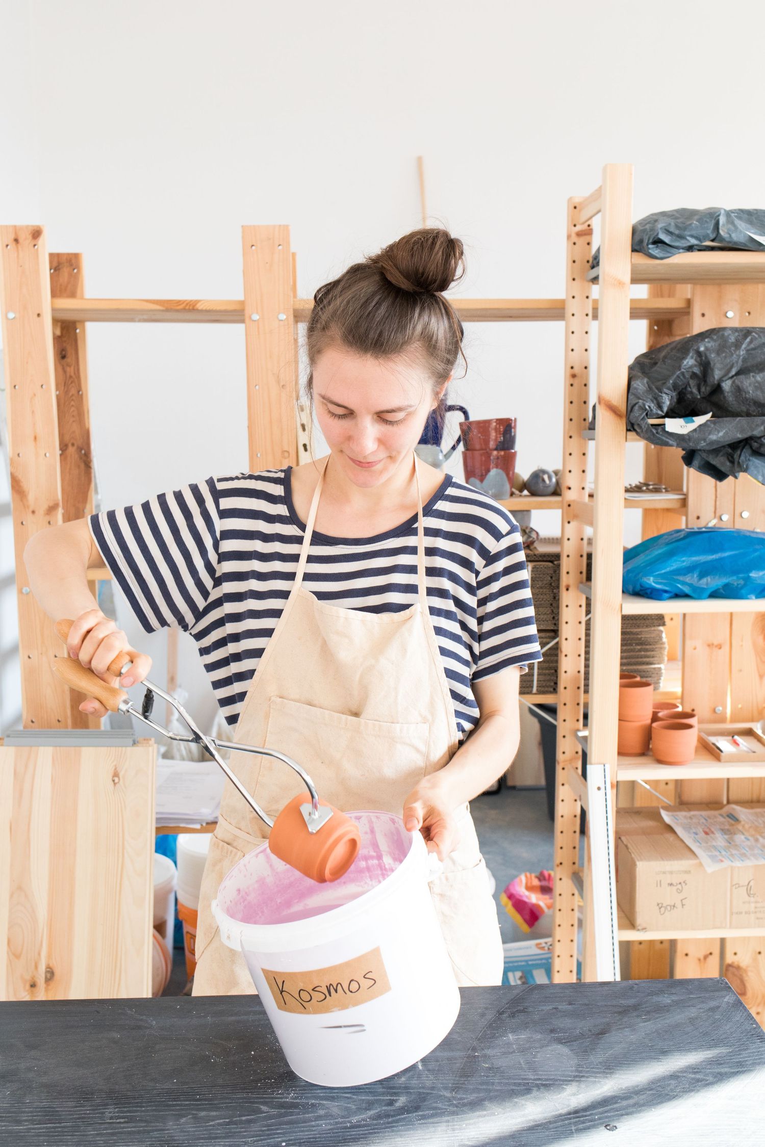 Woman in a striped shirt and apron dips a terracotta pot into a white bucket of pink glaze in a bright pottery studio with wooden shelves.