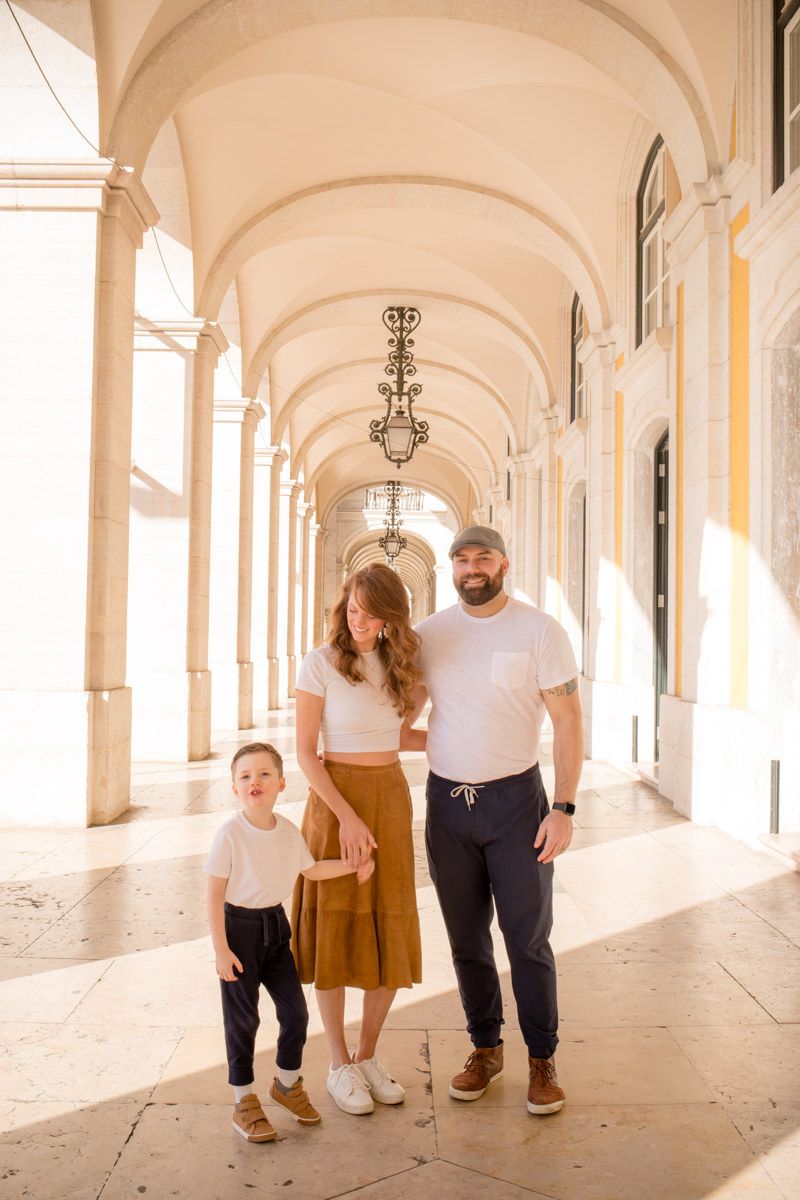 Family photoshoot Lisbon with parents and young son standing beneath a sunlit arched walkway in matching neutral outfits.