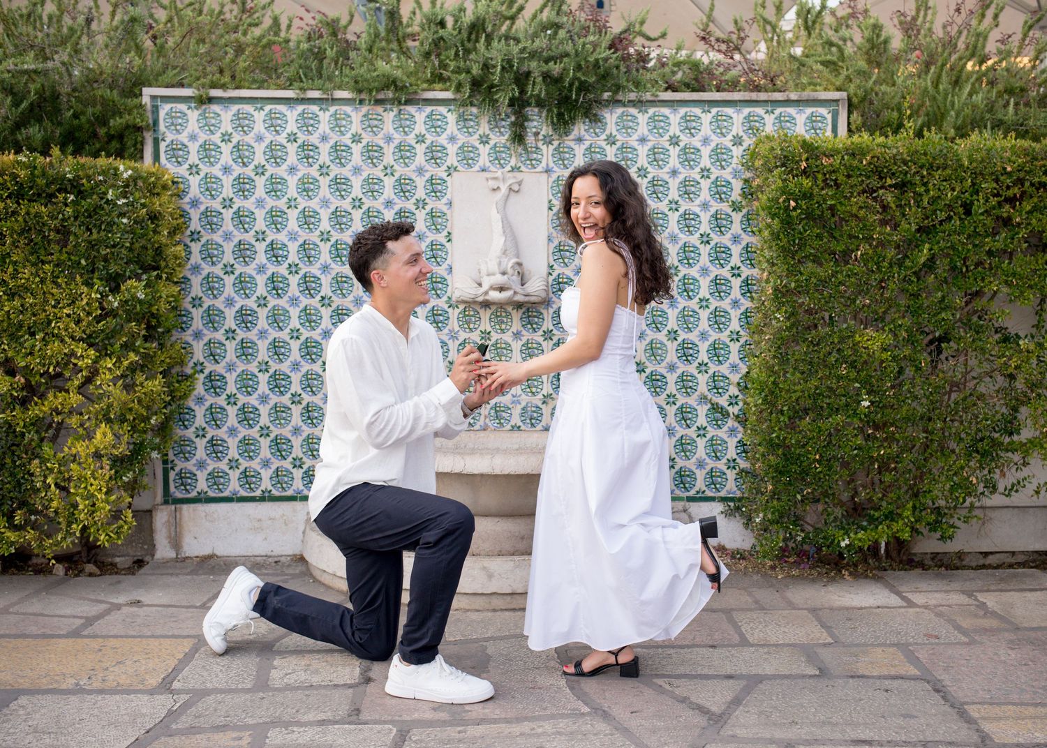 Man kneels proposing to woman in white dress before decorative tiled fountain surrounded by manicured hedges.