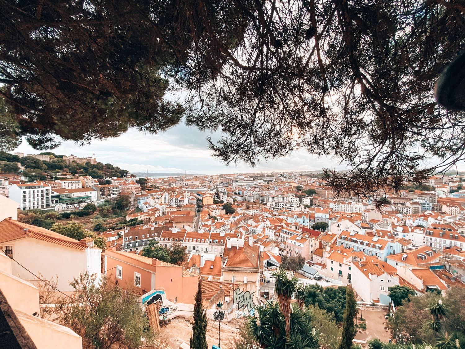 Panoramic view of Lisbon's terracotta rooftops framed by pine trees under a cloudy sky.