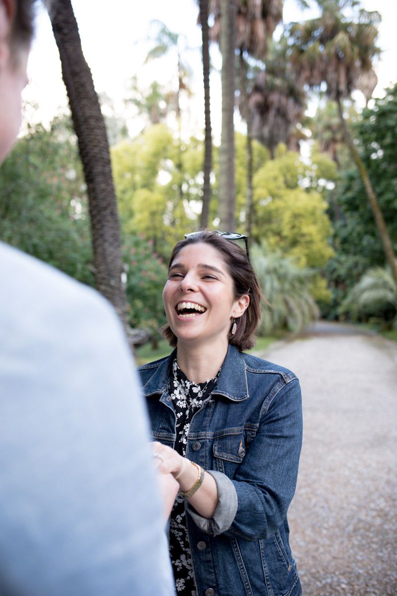 A laughing woman in a denim jacket holds hands with someone on a tree-lined gravel path.