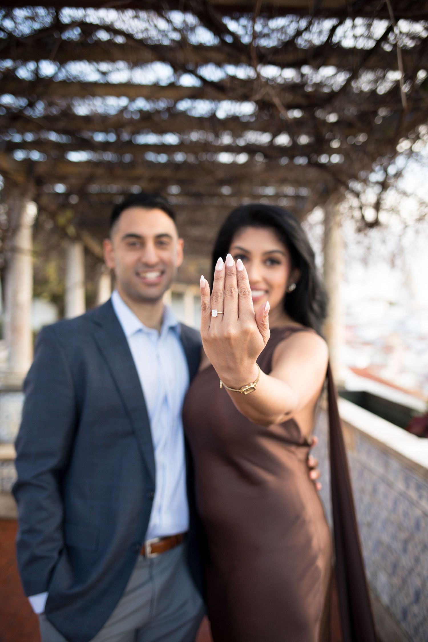 Couple celebrating engagement, woman showing diamond ring toward camera, man in suit smiling, outdoor pergola setting.