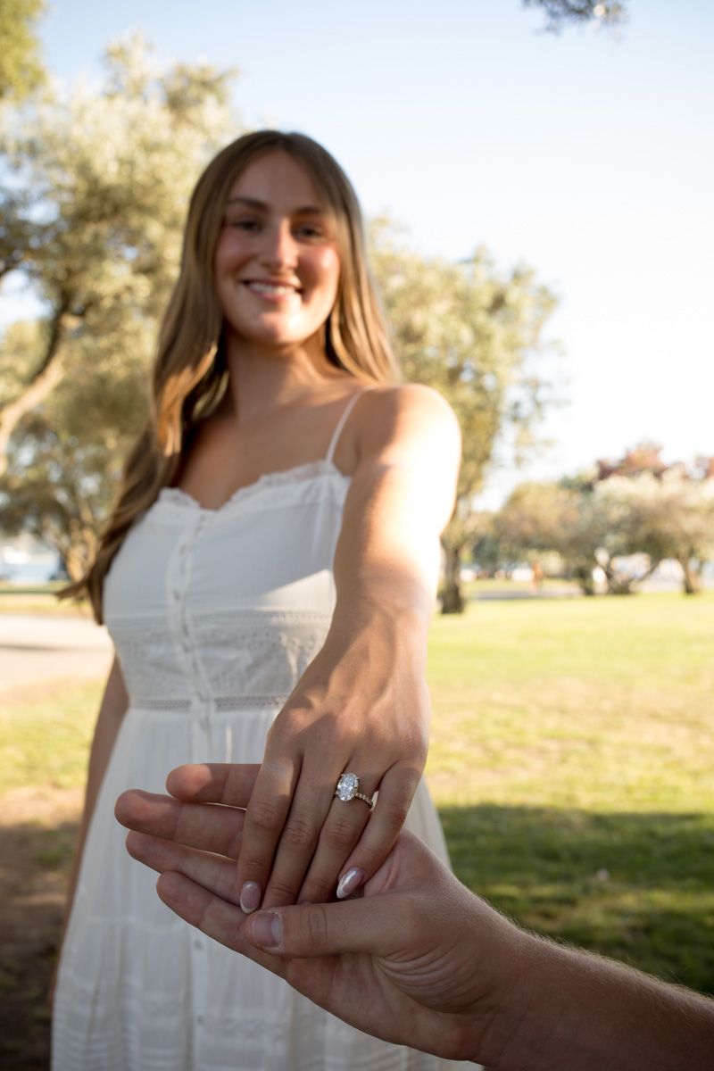 Smiling woman in white dress showing off engagement ring outdoors in sunny park setting.