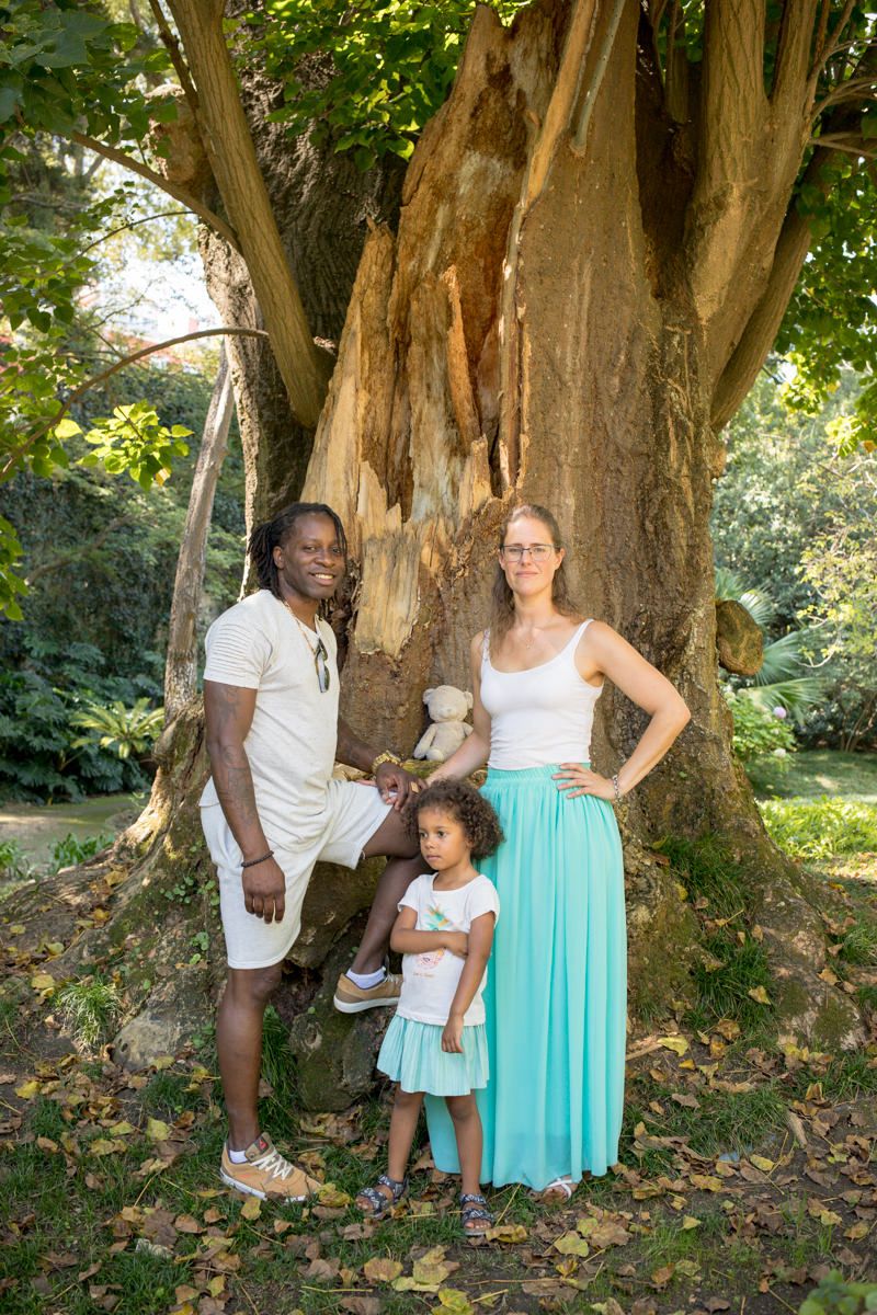 A multiracial family of three poses by a large tree in a lush green park on a sunny day.
