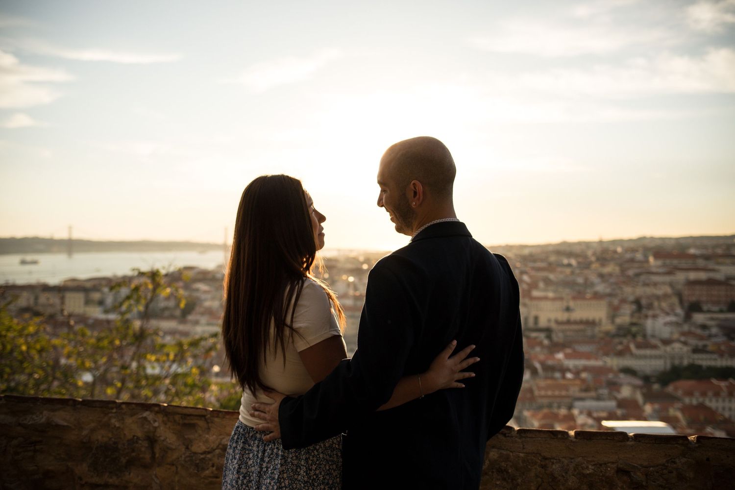 Couple embracing at sunset overlooking Lisbon cityscape and river, romantic silhouette against golden sky.