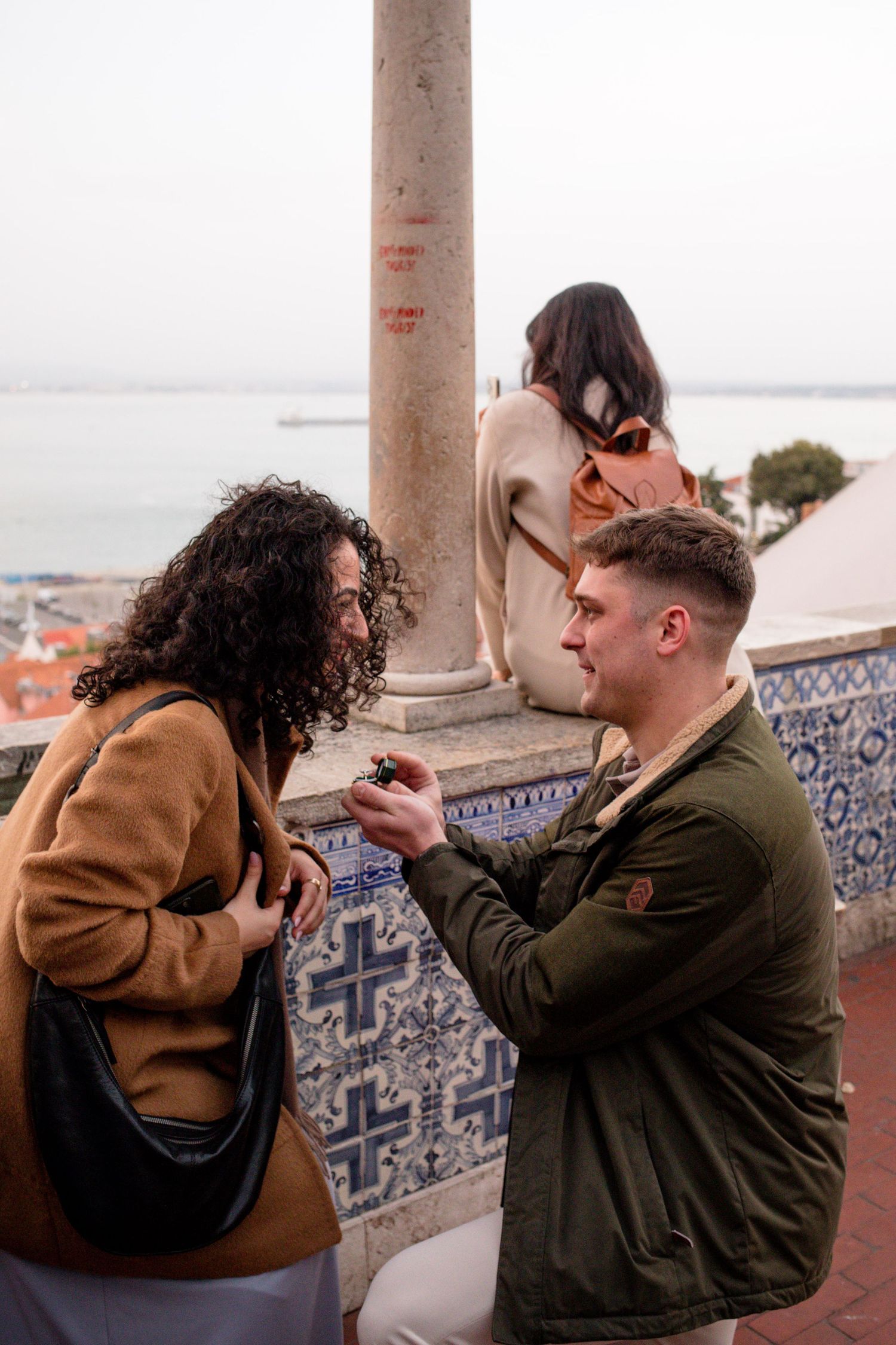Man proposes to woman at scenic overlook with decorative tiles and ocean view in background.