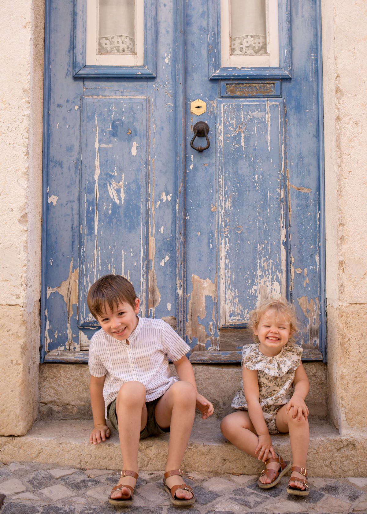 Two happy children sitting on steps in front of a rustic blue peeling painted door, smiling outdoors.