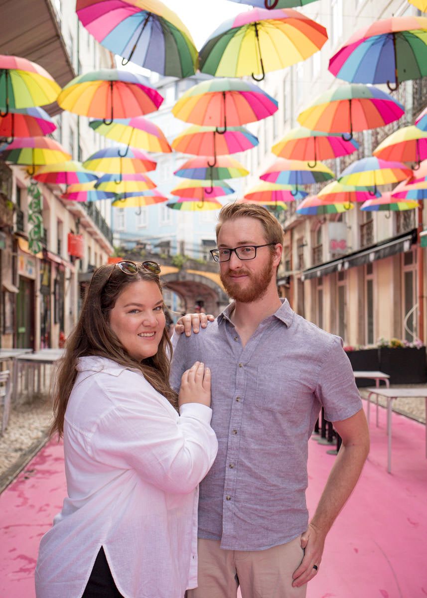 Couple posing under colorful rainbow umbrellas on a pink street in Lisbon, Portugal.