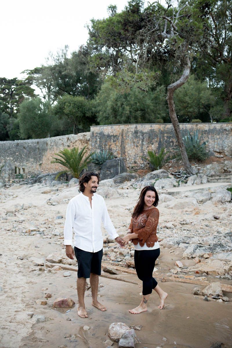 Barefoot couple holding hands walking on rocky beach with lush greenery background.