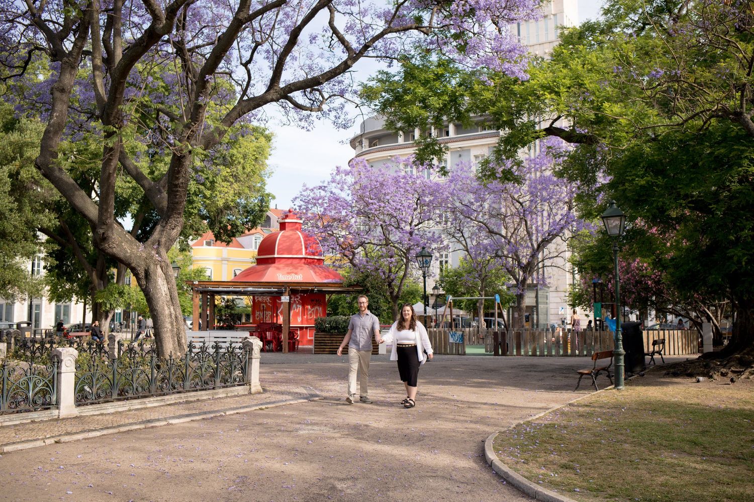 Couple strolling through a scenic park lined with blooming purple jacaranda trees and a red kiosk.