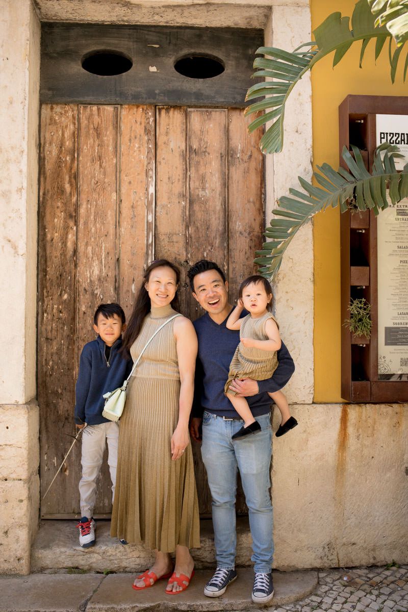 Asian family of four poses before rustic wooden door, father holding toddler while pregnant mother stands with older son.