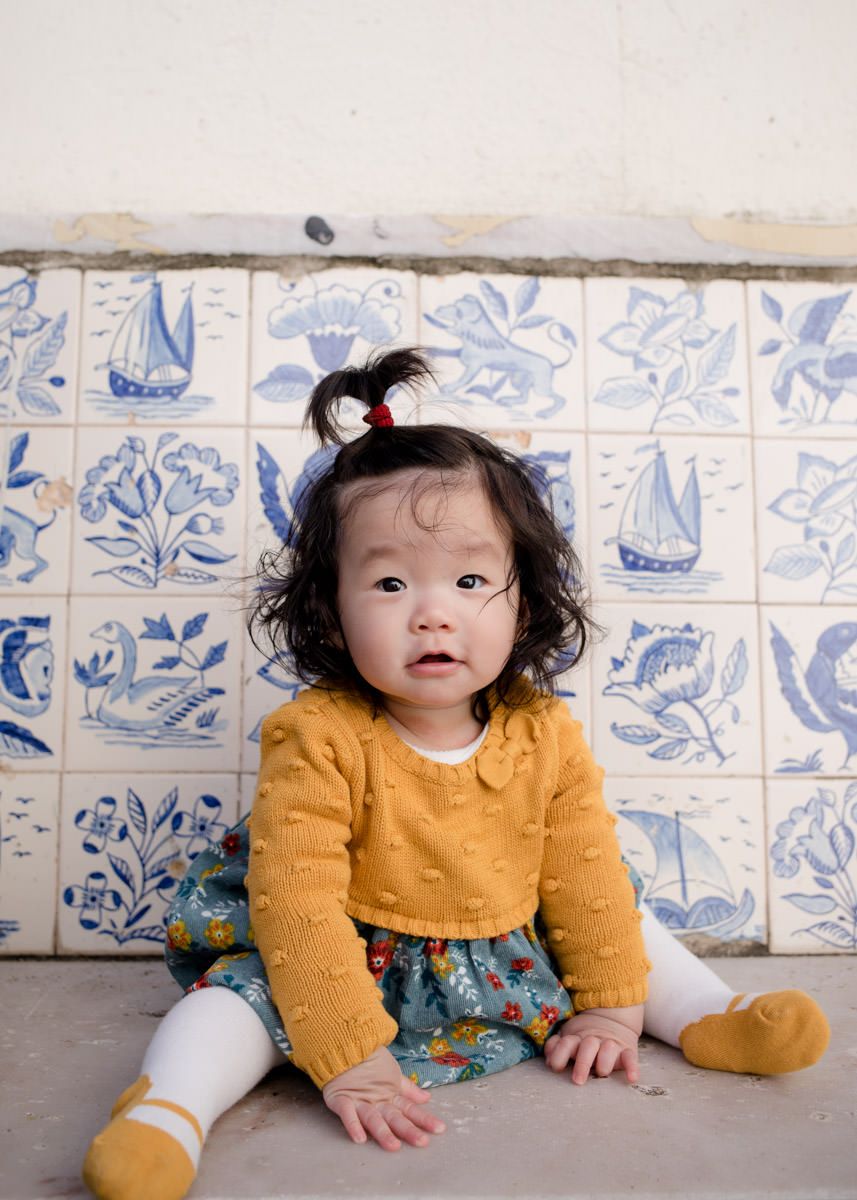 Baby girl in mustard cardigan and floral skirt sitting in front of decorative blue and white tiles, photographed by a Lisbon photographer.