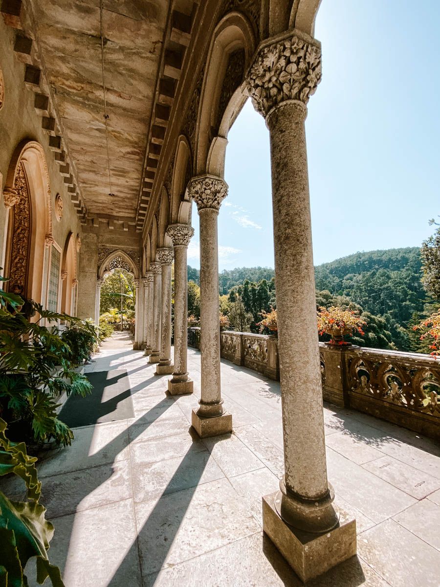 Ornate stone colonnade with decorative capitals at a historic palace, overlooking lush green forest hills under a blue sky.