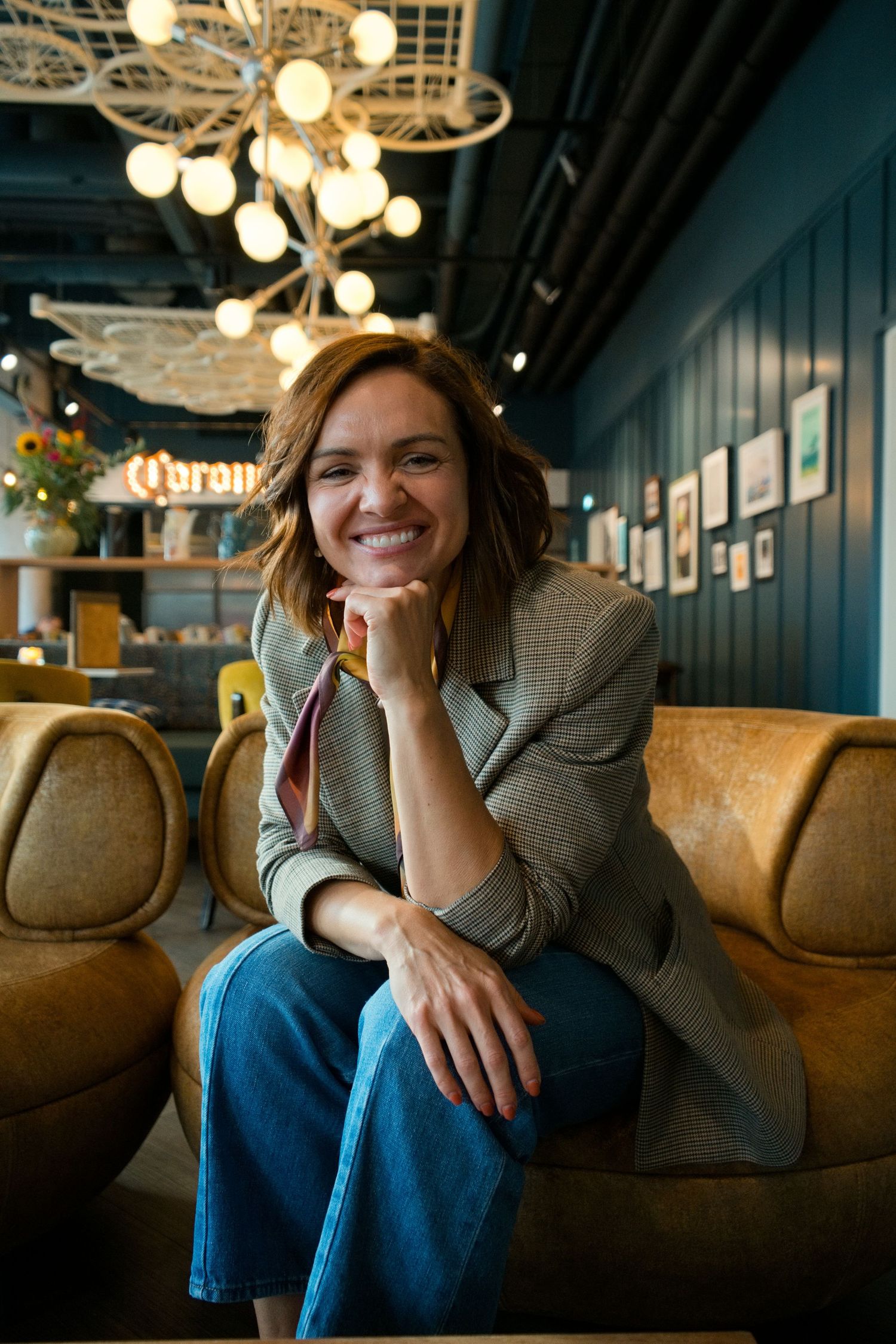 Smiling woman in a plaid blazer and blue jeans sits in a tan leather armchair inside a stylish cafe with modern pendant lights.