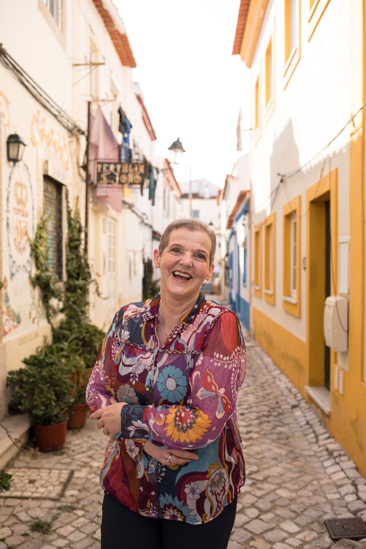 Smiling woman in a colorful floral blouse standing on a cobblestone street lined with white and yellow buildings in a charming old town.