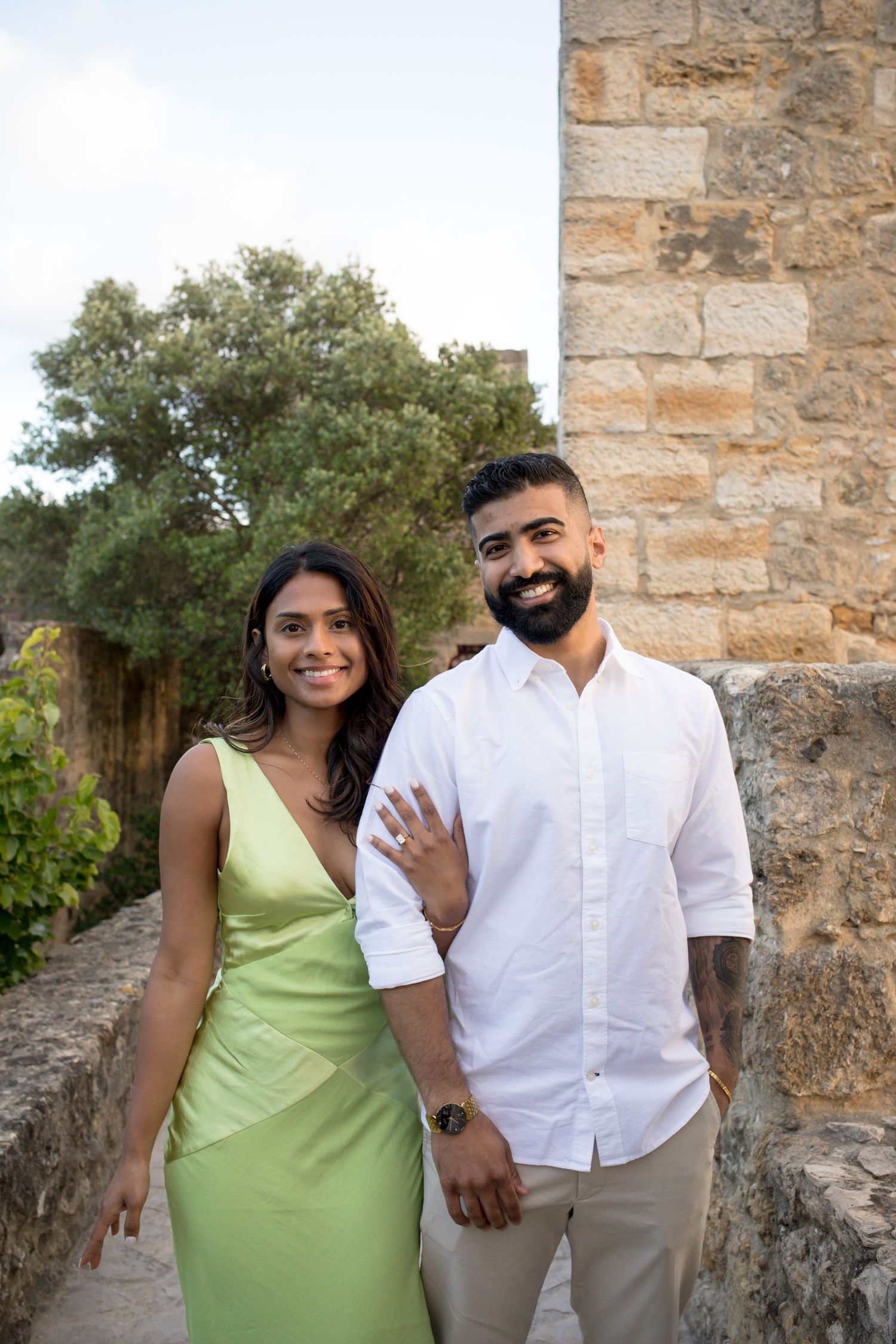Smiling couple posing outdoors near stone wall, woman in green dress, man in white shirt with tattoo sleeve.