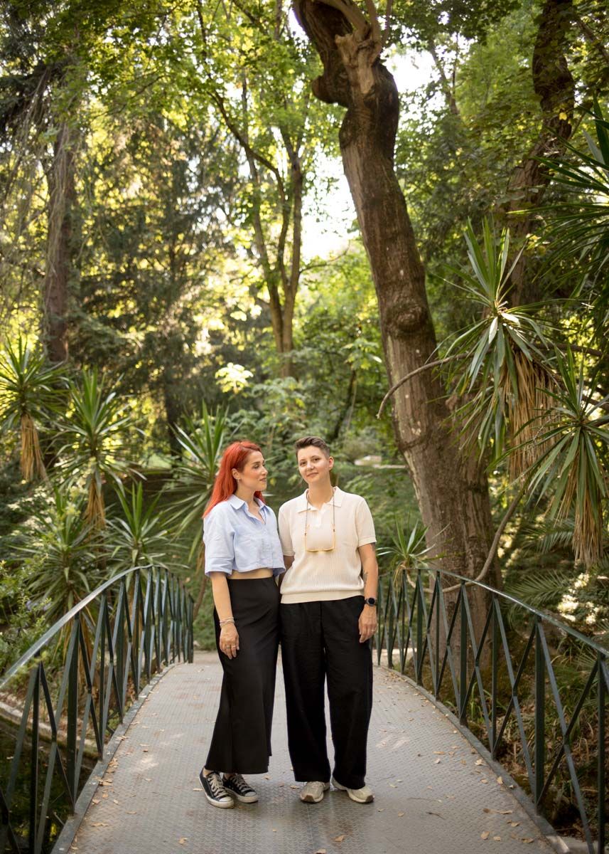 Two women stand on a bridge surrounded by lush green trees in a scenic botanical garden setting.
