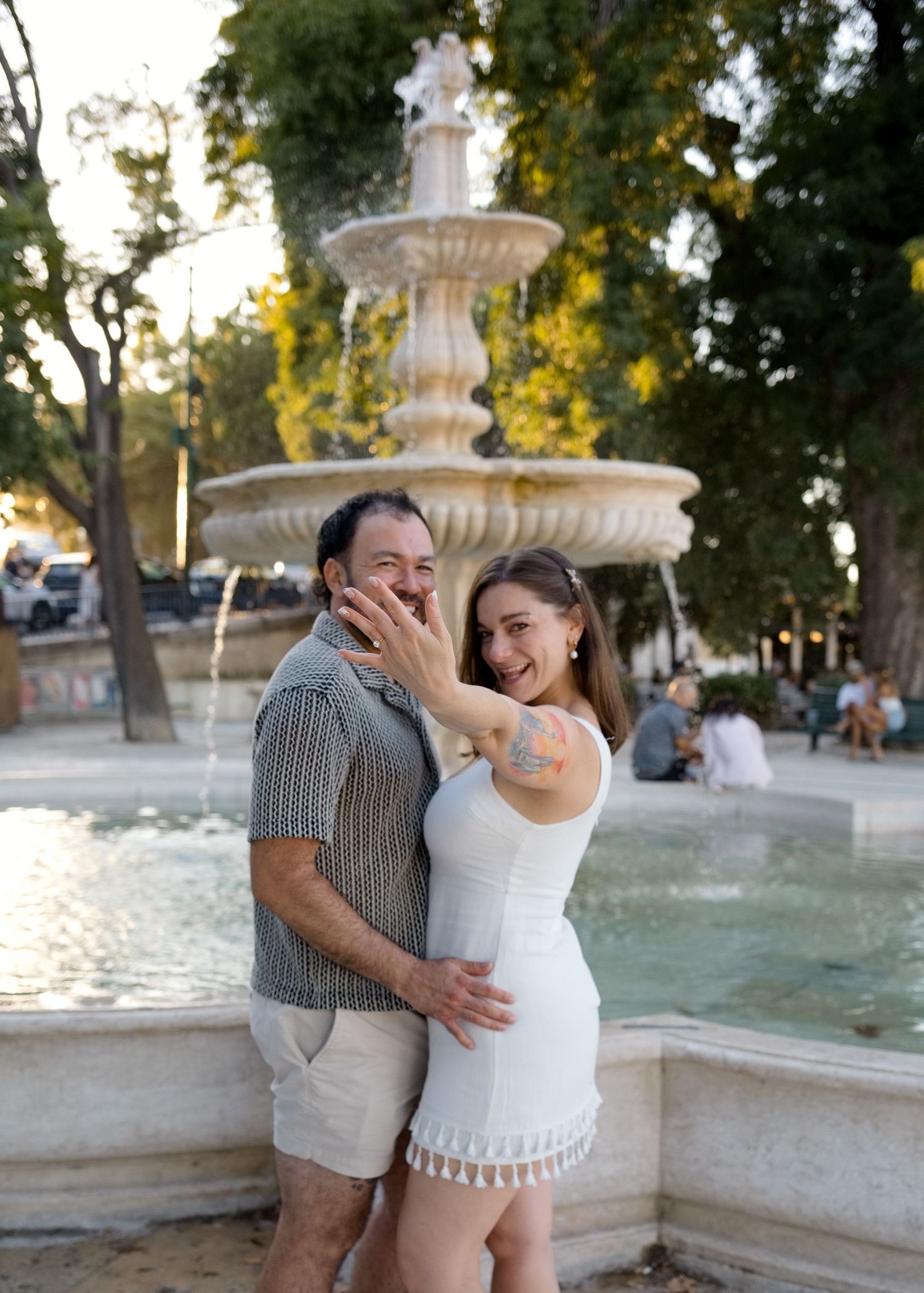 Happy couple embracing and laughing in front of a ornate stone fountain in a sunny park setting.