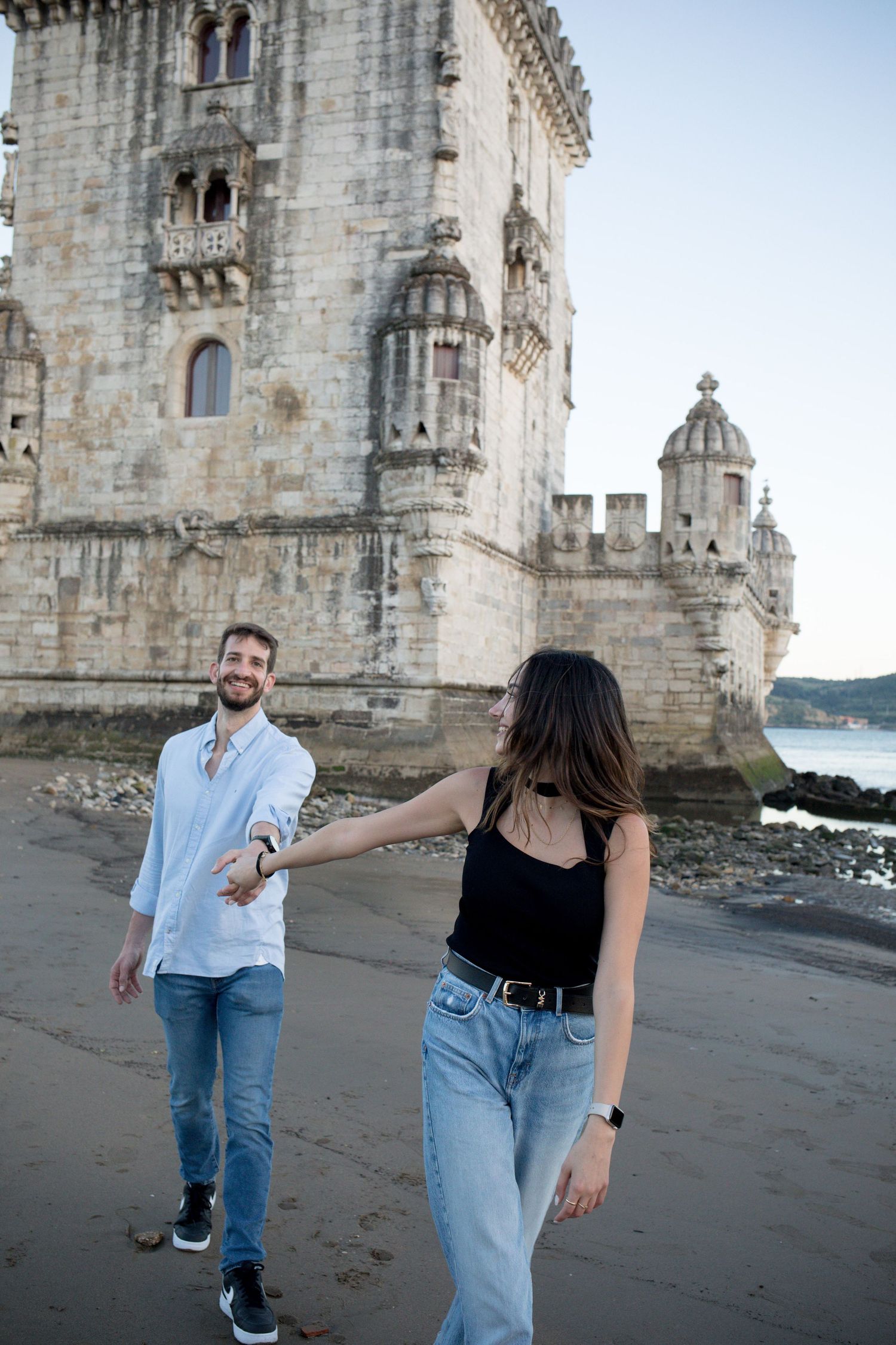 Couple holding hands and walking on the beach near the historic Belem Tower in Lisbon, Portugal.