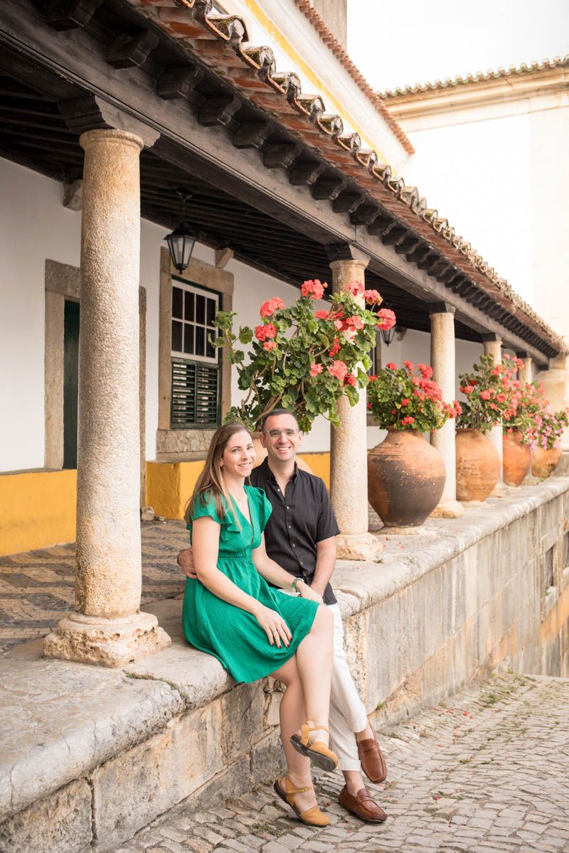 Couple sitting together on a stone ledge beside colorful geraniums and columns of a traditional Portuguese building.