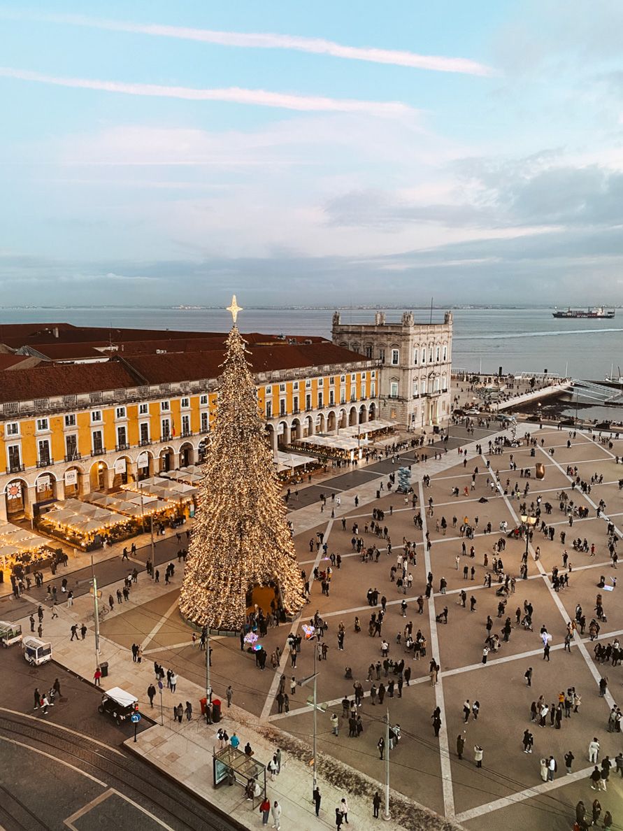 Aerial view of Lisbon's Praça do Comércio with a large Christmas tree, yellow buildings, and the Tagus River at dusk.