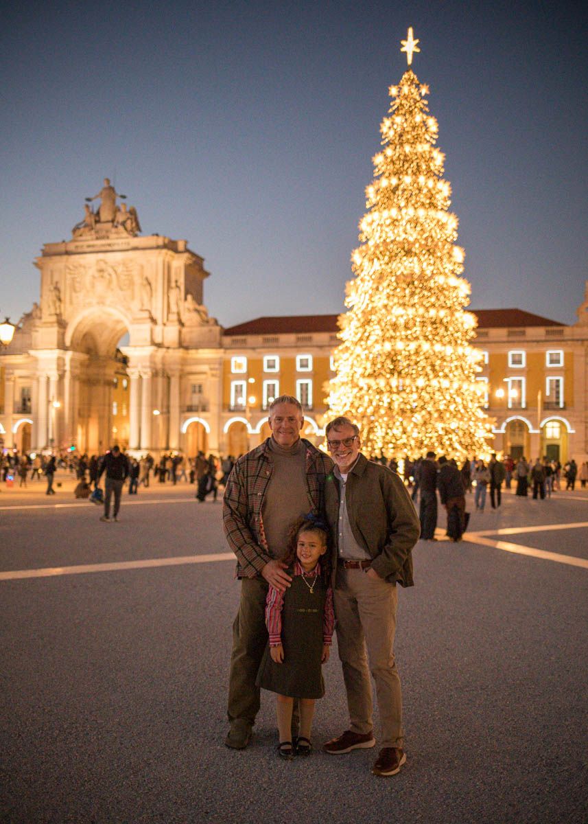 Two men and a child pose in front of a tall illuminated Christmas tree at Lisbon's Praca do Comercio at dusk.