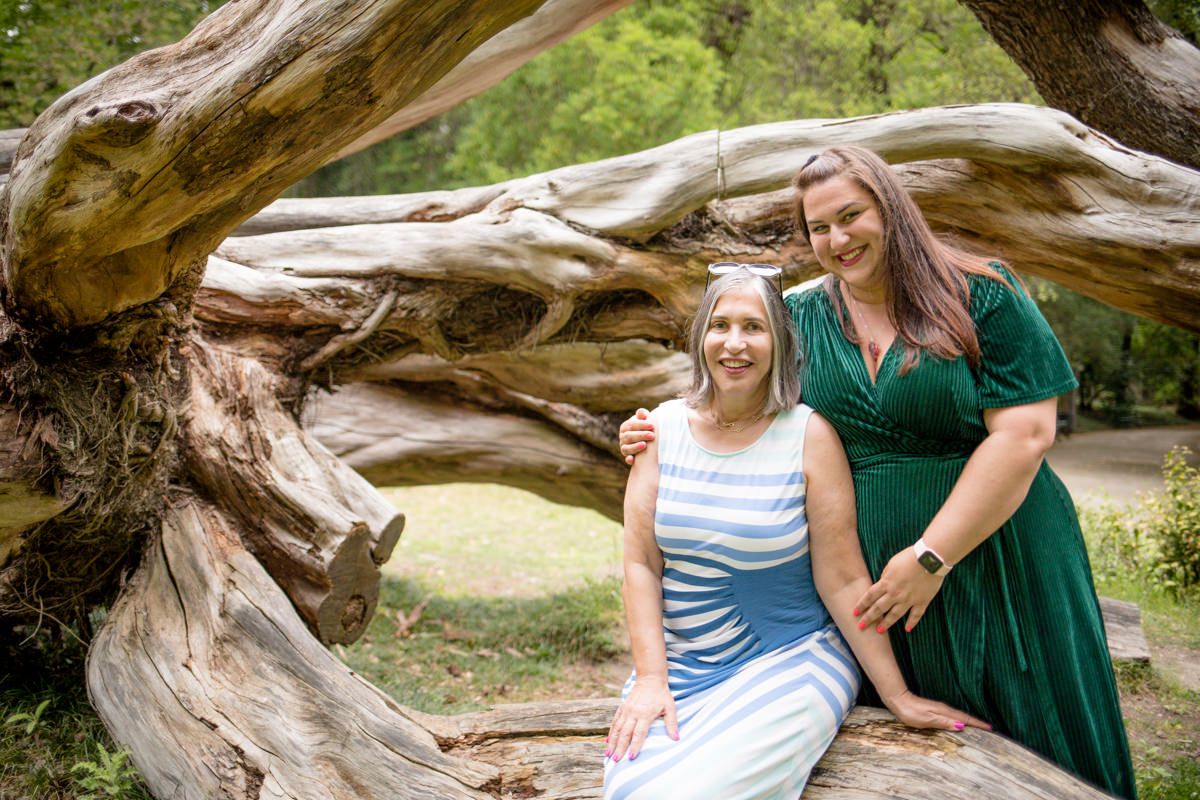 Two women pose together on a large twisted tree trunk in a lush green park, one in green dress, one in striped blue.