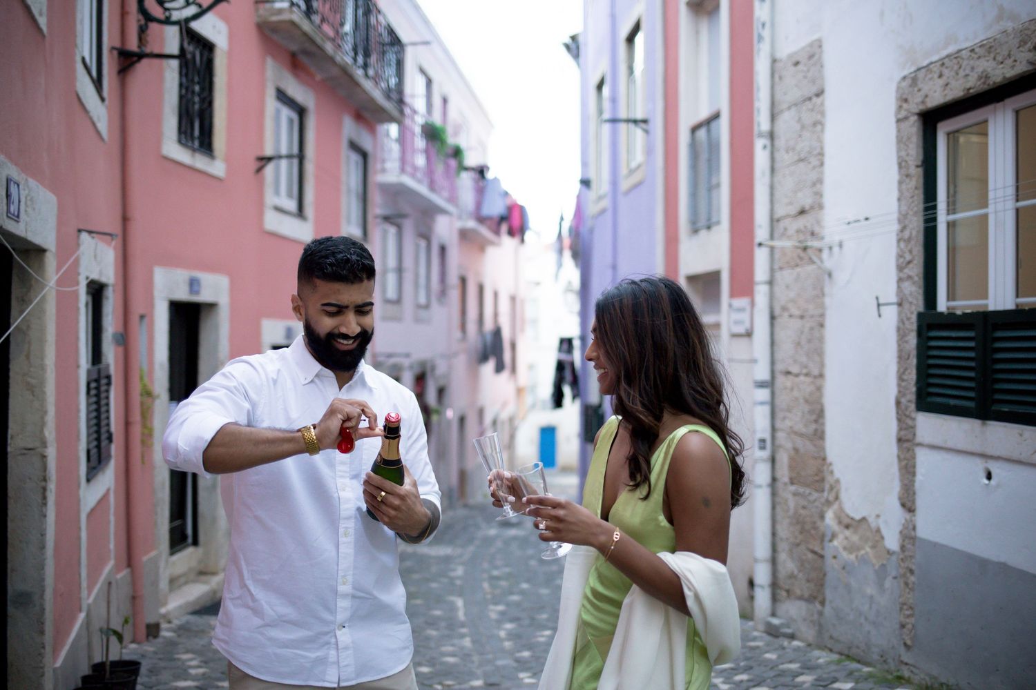 Couple enjoying drinks on a colorful European street, man opening a beer bottle while woman holds a glass.