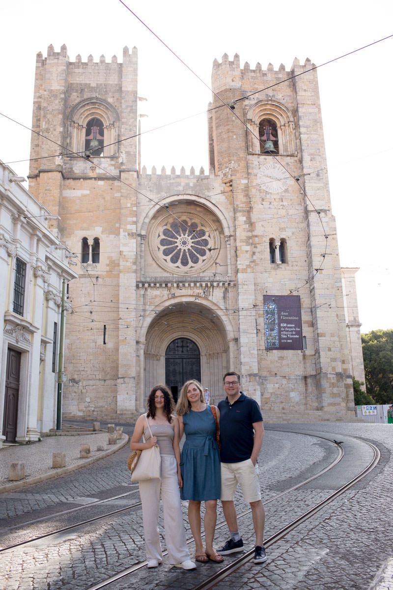 Three tourists posing in front of Lisbon Cathedral, Portugal, with tram tracks on cobblestone street.
