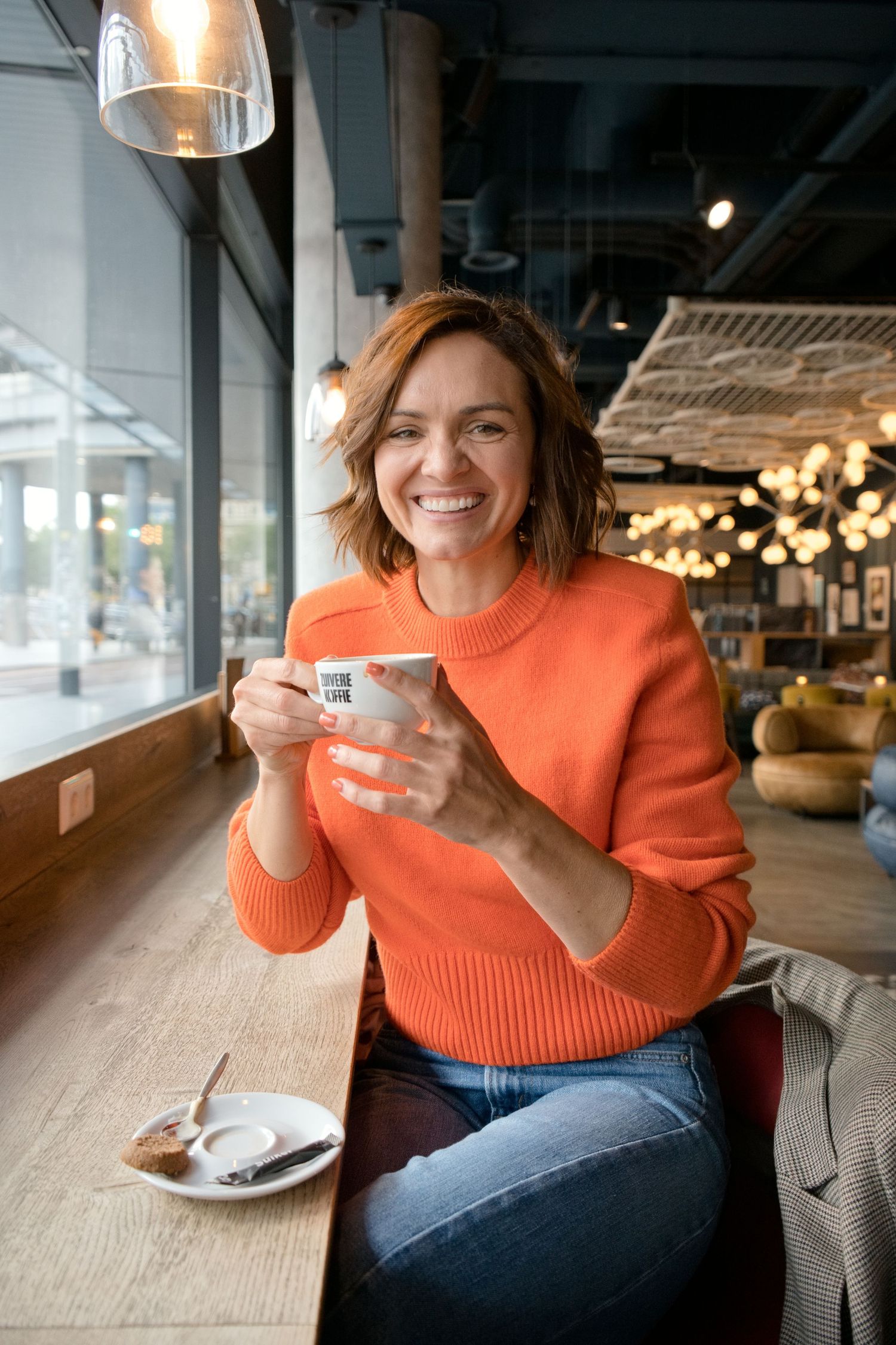 Smiling woman in an orange sweater holds a white espresso cup at a wooden window counter in a cozy cafe with warm string lights.