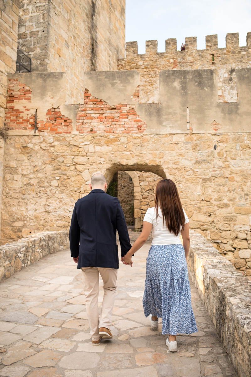 Couple walking hand in hand through ancient stone castle archway on cobblestone path.