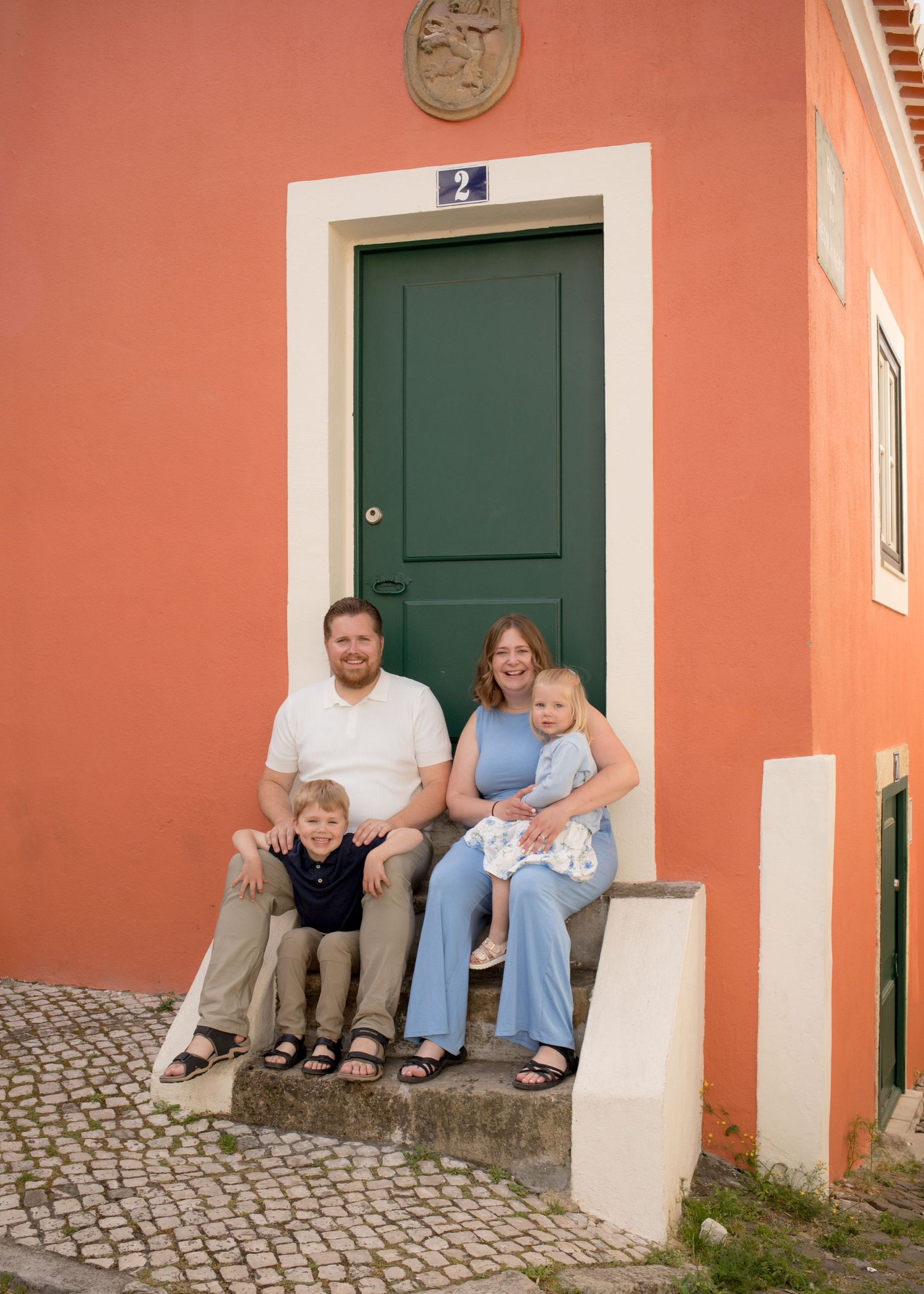 Young family of four sitting on steps of an orange building with green door in Portugal.