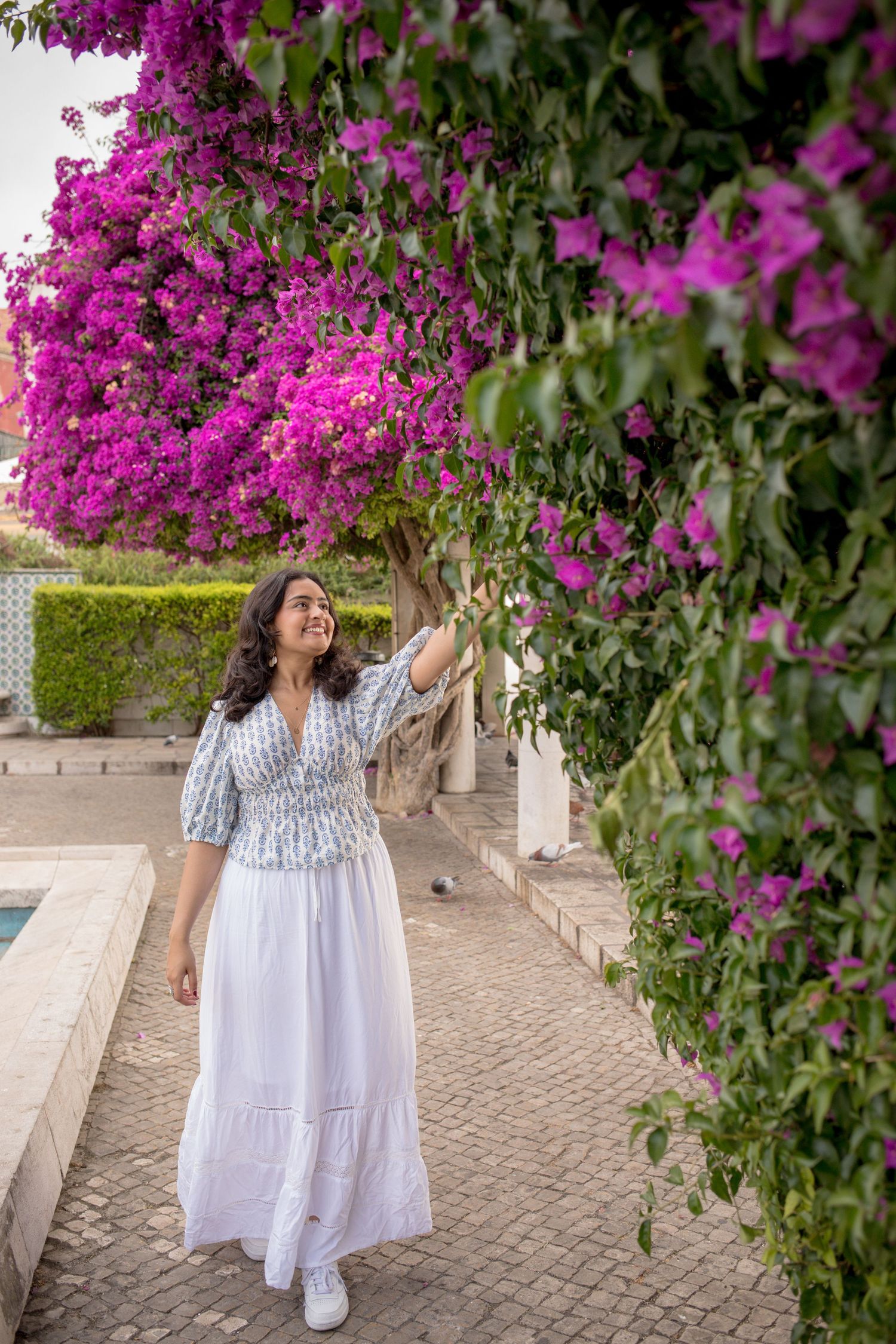 Woman in a white skirt reaching up to touch vibrant pink bougainvillea flowers outdoors.
