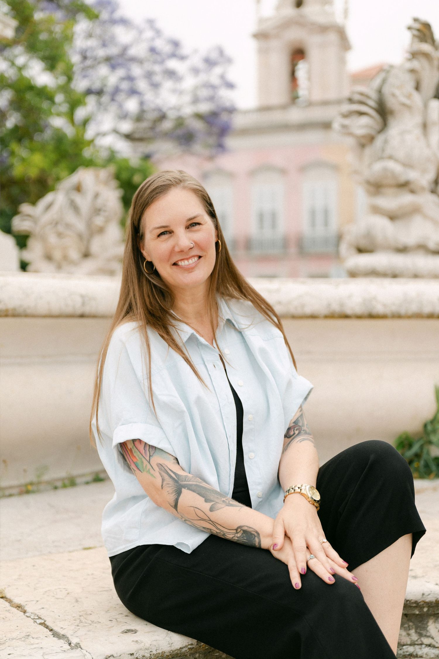 Smiling woman with tattoos sits on stone fountain ledge wearing white shirt and black pants in a Lisbon plaza.
