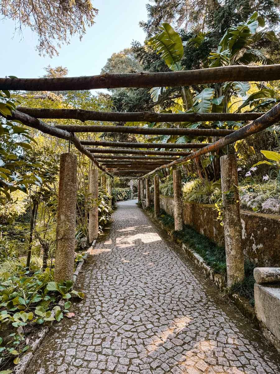 A rustic cobblestone pathway lined with wooden pergola columns and lush tropical greenery in a serene garden.