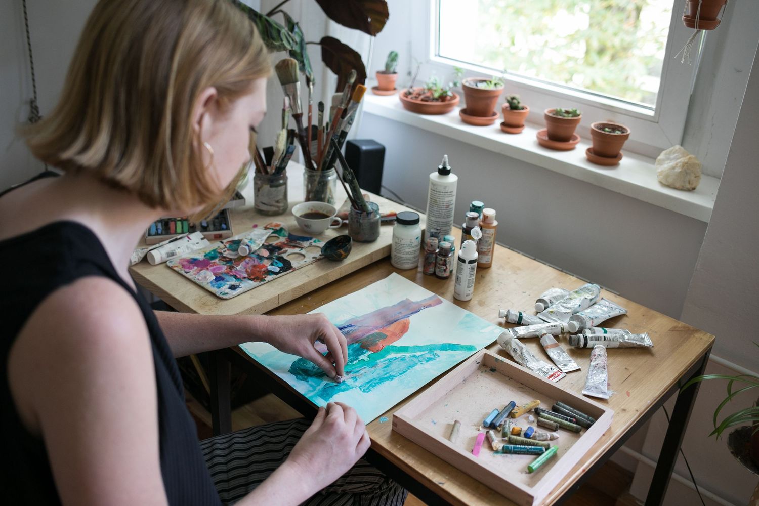 Woman with blonde hair painting a colorful watercolor artwork at a wooden desk with brushes, paint tubes and plants by a sunny window.