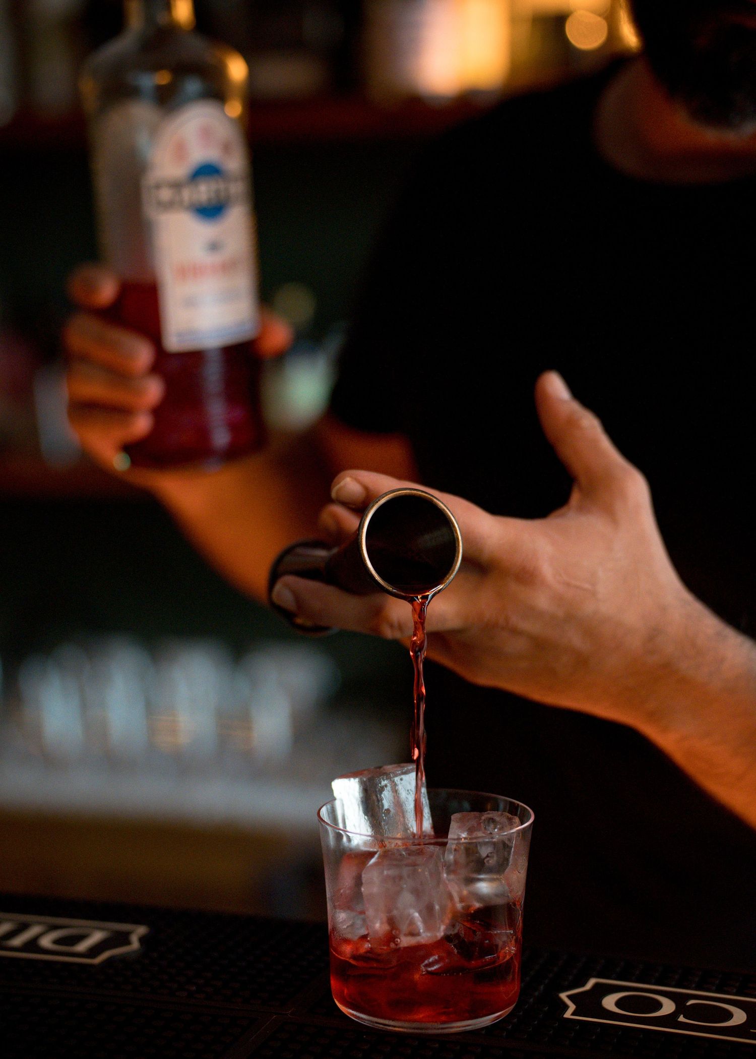 Bartender pouring a red cocktail from a jigger into a rocks glass filled with ice at a dimly lit bar.