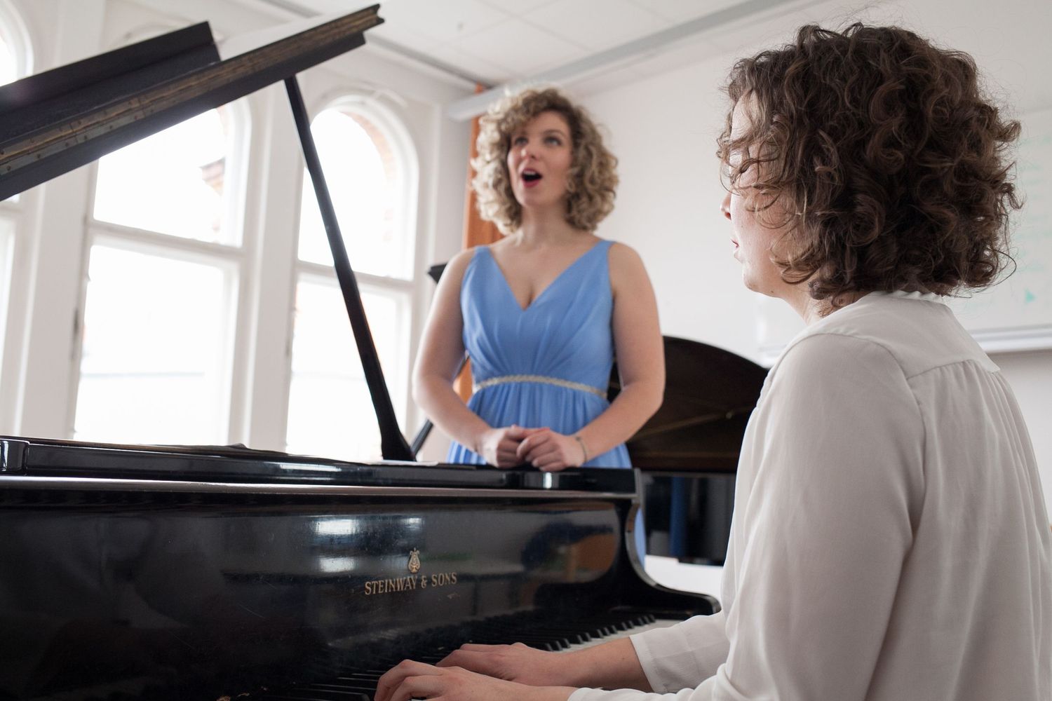 A woman in a blue dress sings in a bright room while a curly-haired pianist plays a black grand piano in the foreground.